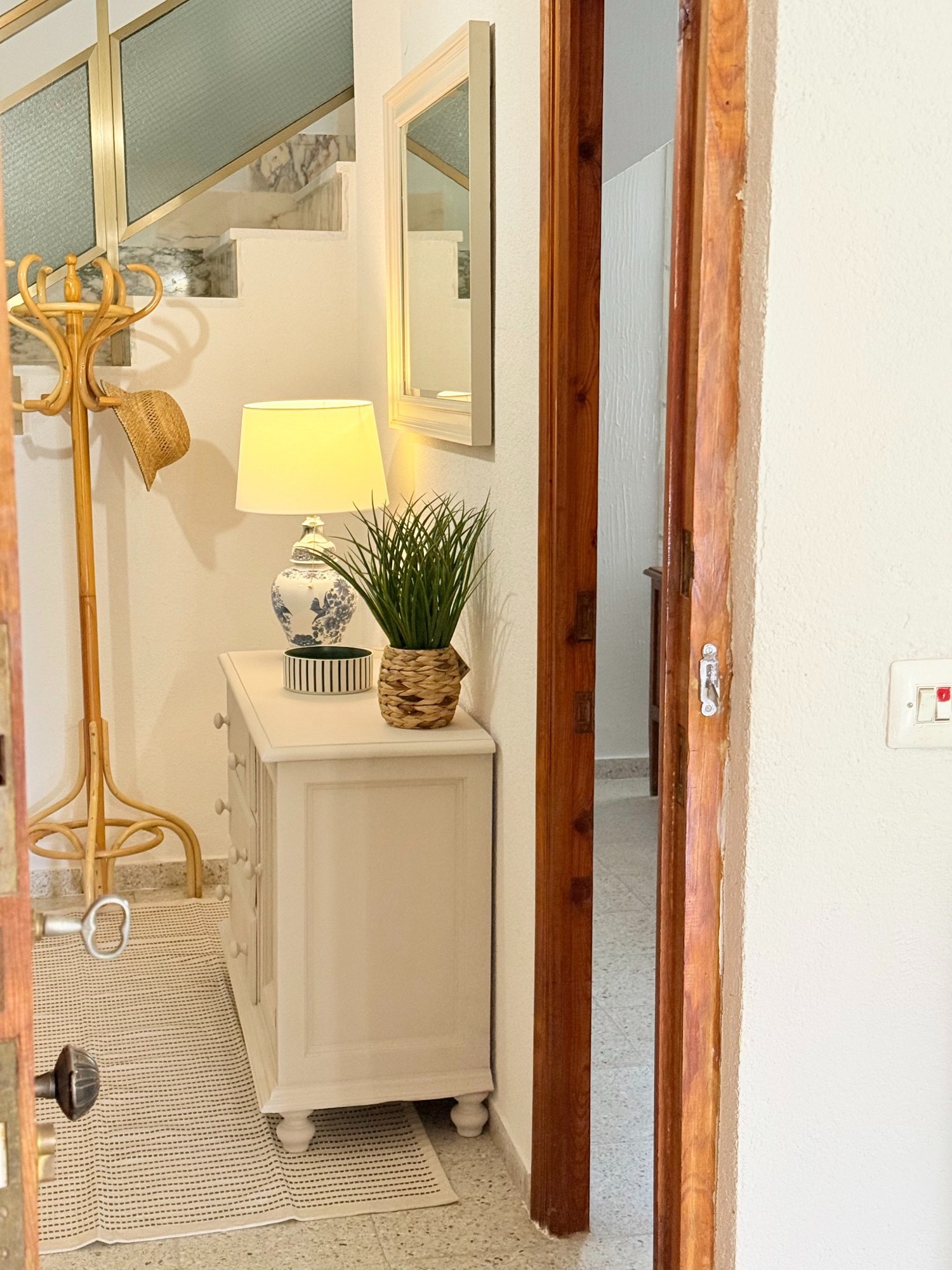 Después: entrada vista desde la puerta, con mueble claro, lámpara encendida, planta y perchero